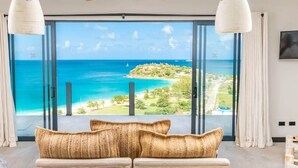 A modern living room with a large window overlooking a beach and ocean. - Panorama Suite – Oceanview Luxury Suite at Tamarind Hills, Antigua (Unknown)