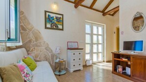 A cozy living room with a stone wall, a white sofa with colorful cushions, a small side table, a TV on a wooden stand, and a large window with a view. - Dubrovnik Chalets (Konavle)