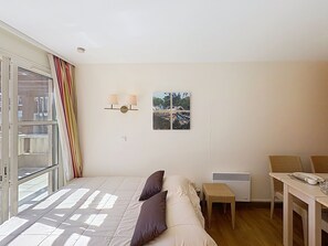 Un dormitorio con cama, ventana con cortinas, lámpara de pared, un cuadro, una mesita y una silla.