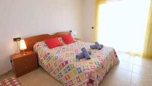 A neatly made bed with a colorful geometric-patterned duvet, a wooden nightstand with a lamp, and a small bench with a plaid cushion. - Golden Beach by Interhome (St Carles de la Ràpita)