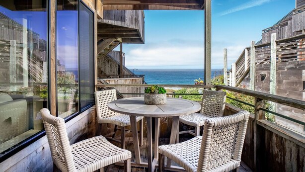 Terraza de madera con sillas de mimbre y una mesa redonda, con vistas a la costa.
