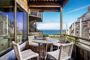 Eine Holzterrasse mit Korbstühlen und einem runden Tisch, mit Blick auf die Küste.