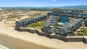Un hotel frente al mar con varias construcciones, una playa y montañas al fondo.