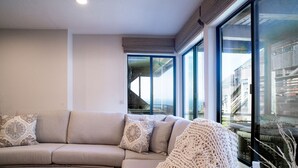 Una sala de estar moderna con un sofá seccional color beige, una manta tejida y grandes ventanales con vistas a la ciudad.