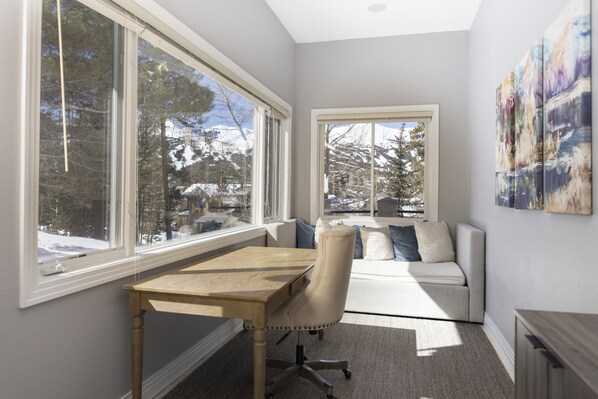A room with a desk, a cushioned chair, a sofa, and large windows offering a view of snow-covered trees.