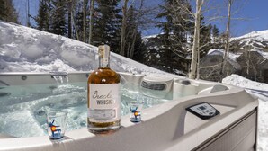 Un jacuzzi con Breckenridge Whiskey y dos vasos para shots, rodeado de nieve y árboles.