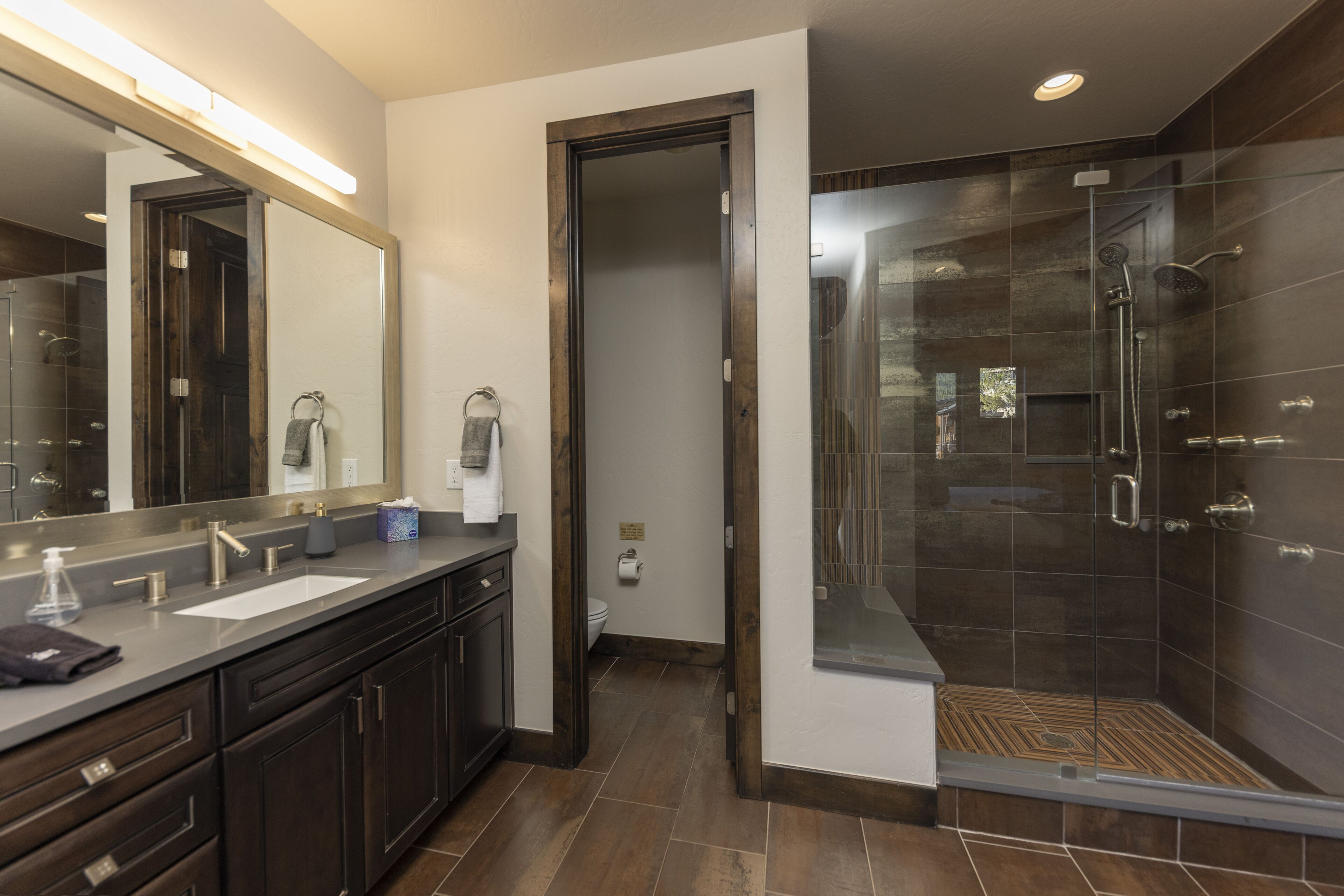 A modern bathroom with a large mirror, dual sinks, a shower enclosure, and dark wood cabinetry.