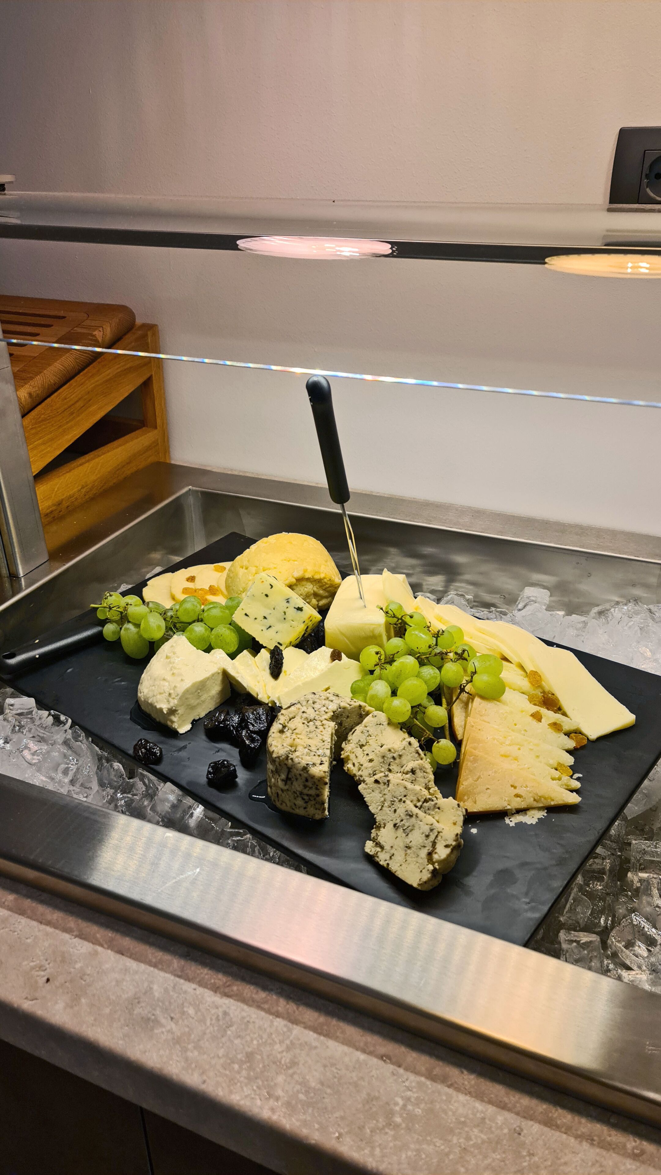 A cheese and fruit platter on a black slate with a knife, ice, and a wooden cutting board in the background.