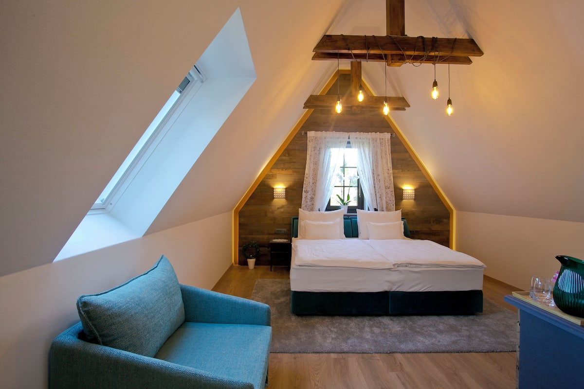A bedroom with a sloped ceiling, a bed, a blue chair, and a small table with a vase.