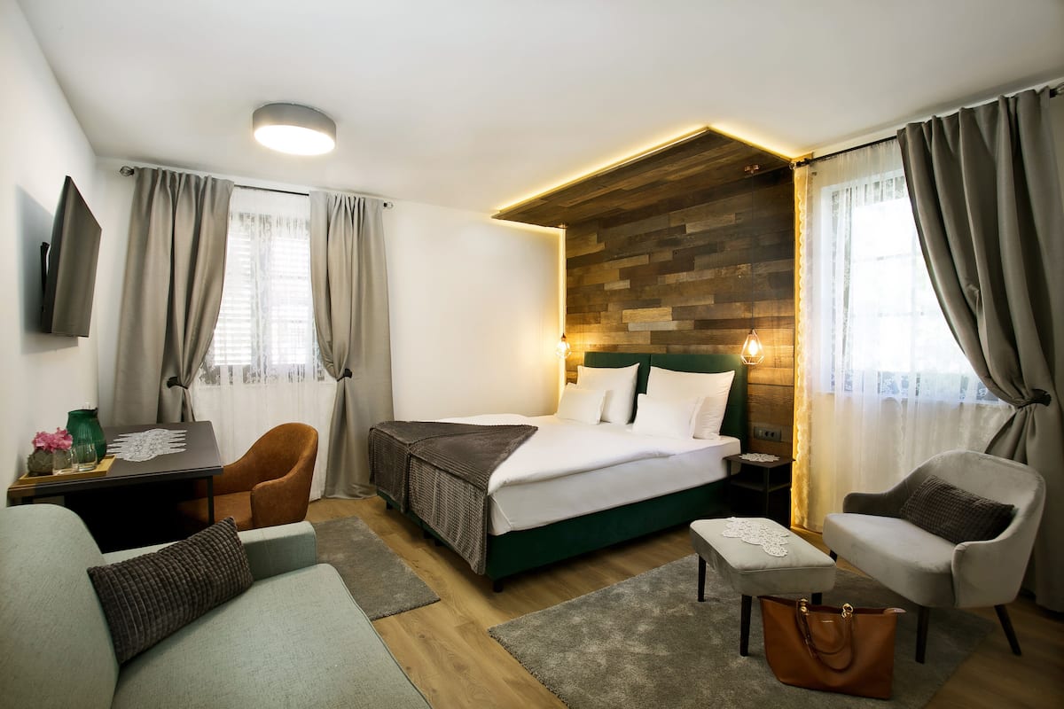 A modern hotel room with a wooden accent wall, a bed with white linens, a desk with a laptop, a chair, a sofa, and a small table with a vase.