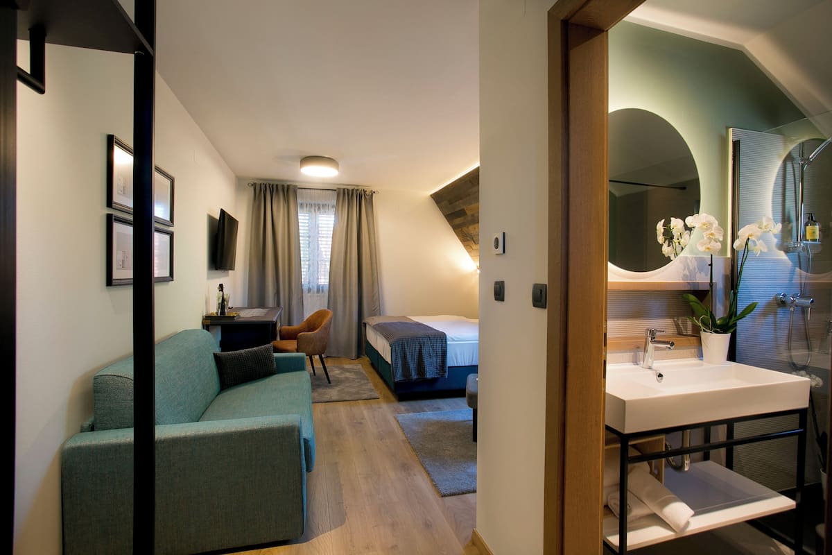 A modern hotel room with a sofa, a bed, a desk, and a bathroom with a sink and mirror.