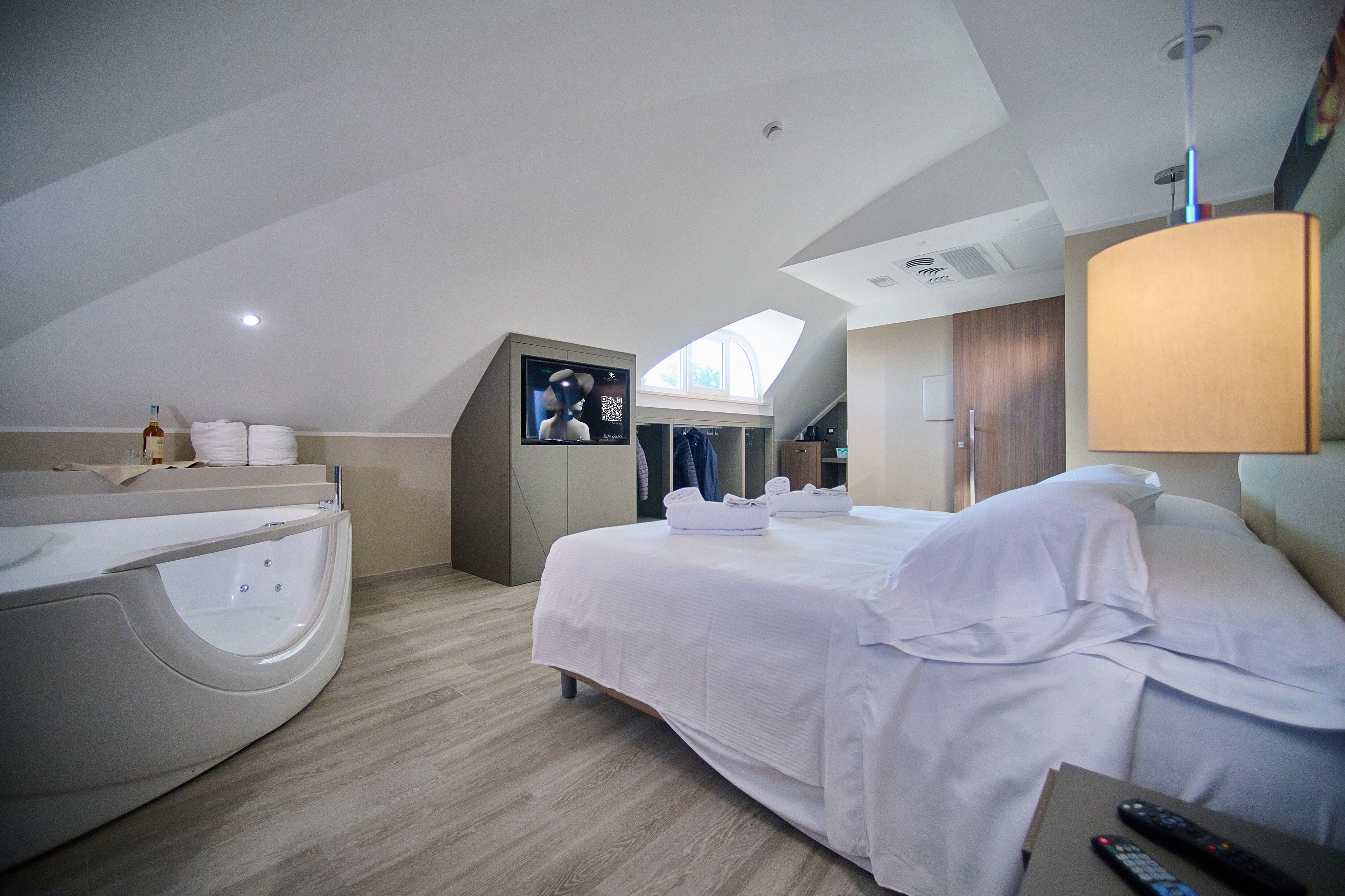 A modern hotel room with a large bed, a built-in bathtub, a TV, and a skylight.