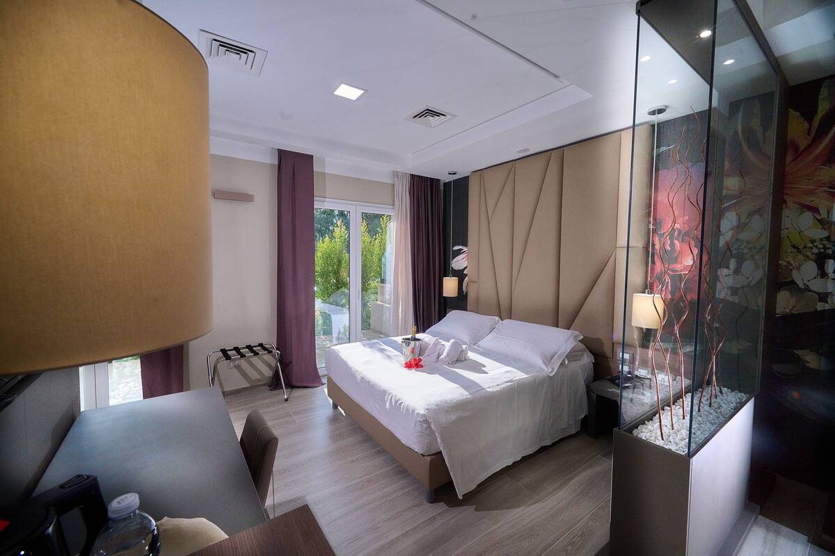 A hotel room with a bed, bedside table, lamp, and a view of greenery outside.