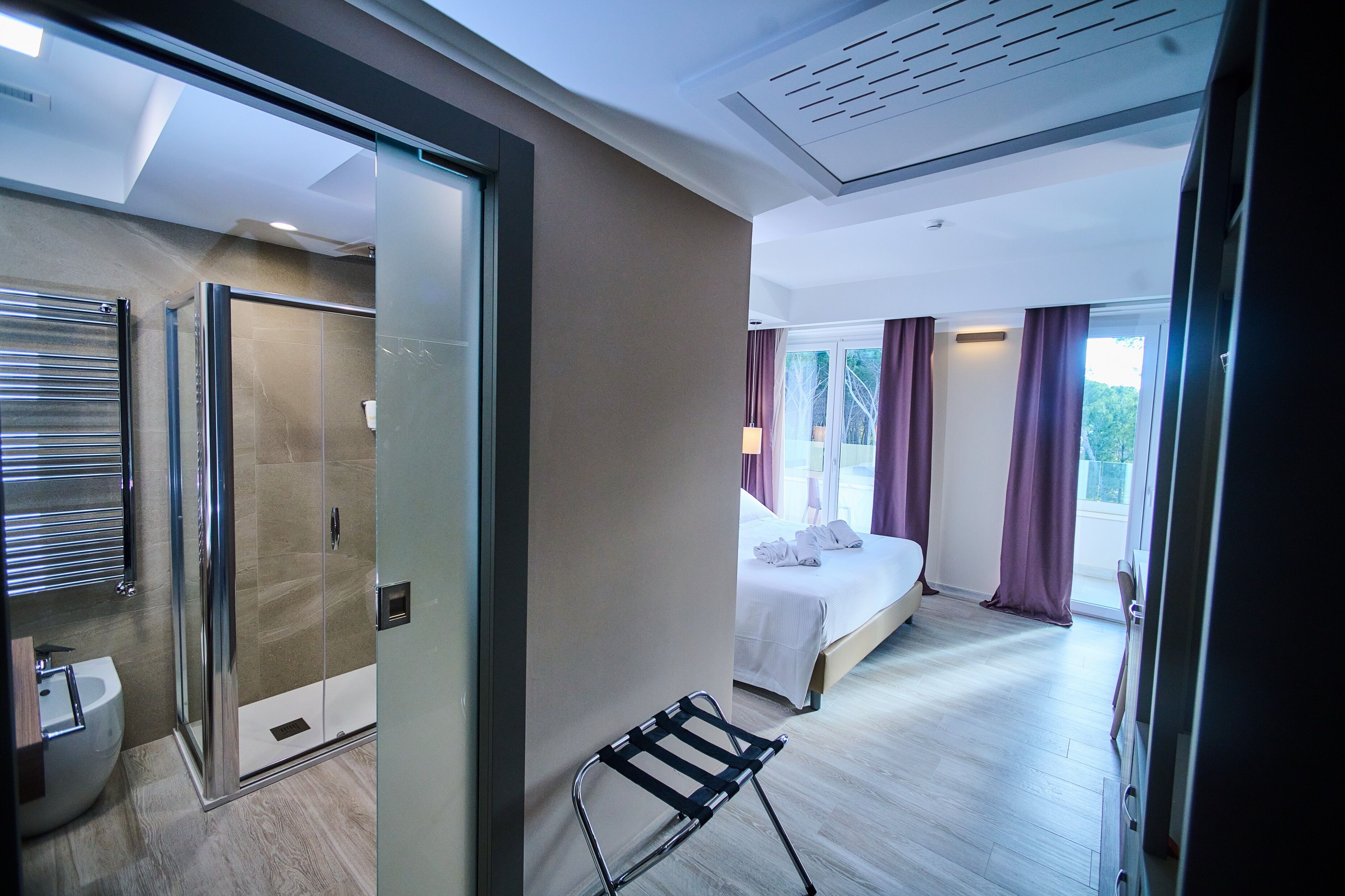 Habitación de hotel moderna con cabina de ducha de vidrio, mesita de noche, cama con ropa blanca y vistas al exterior a través de amplios ventanales.