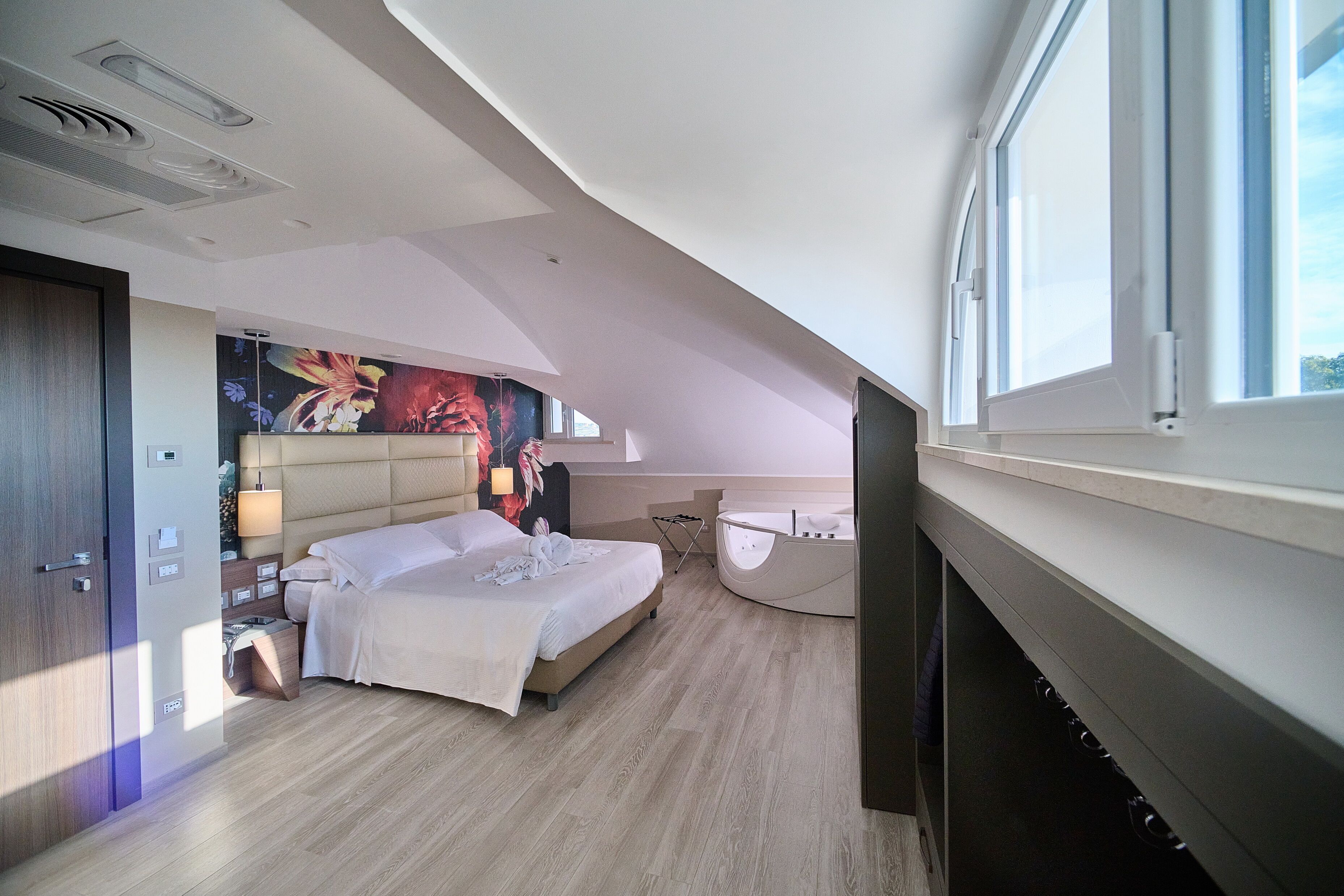 A modern bedroom with a large bed, a bedside table, a nightstand, a wall-mounted TV, and a window with a view of the sky.