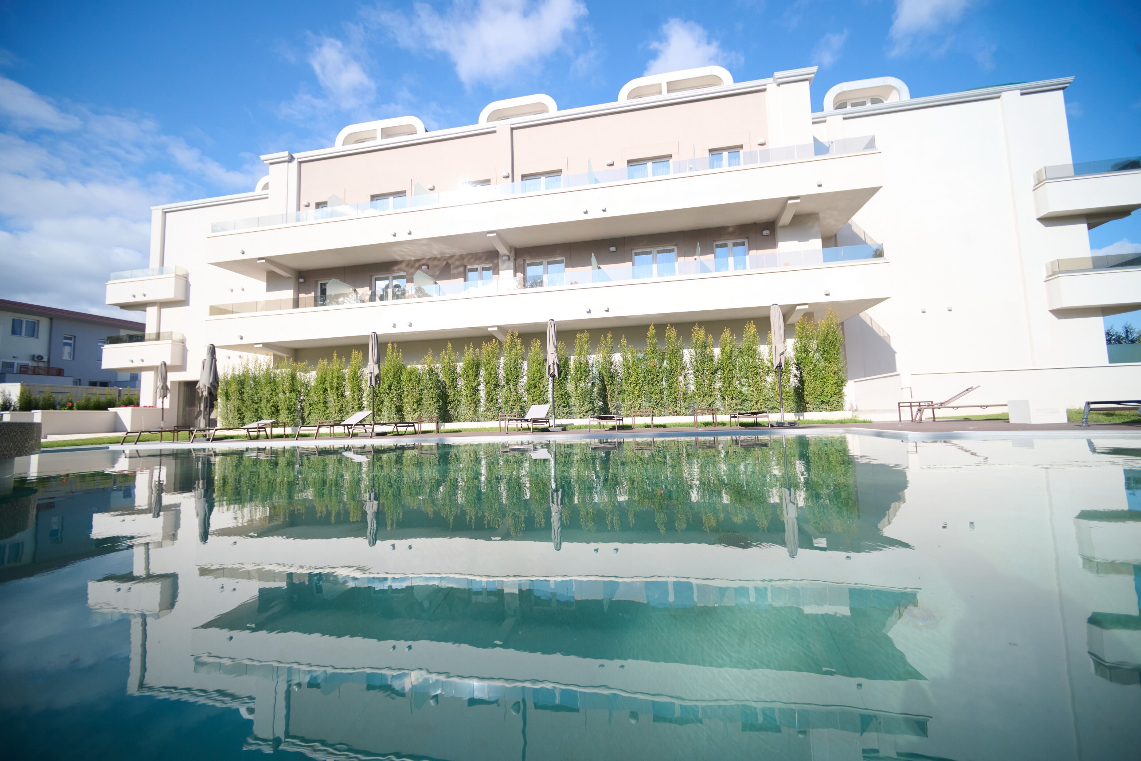 A modern hotel with a large pool, multiple balconies, and a reflection of the building in the water.