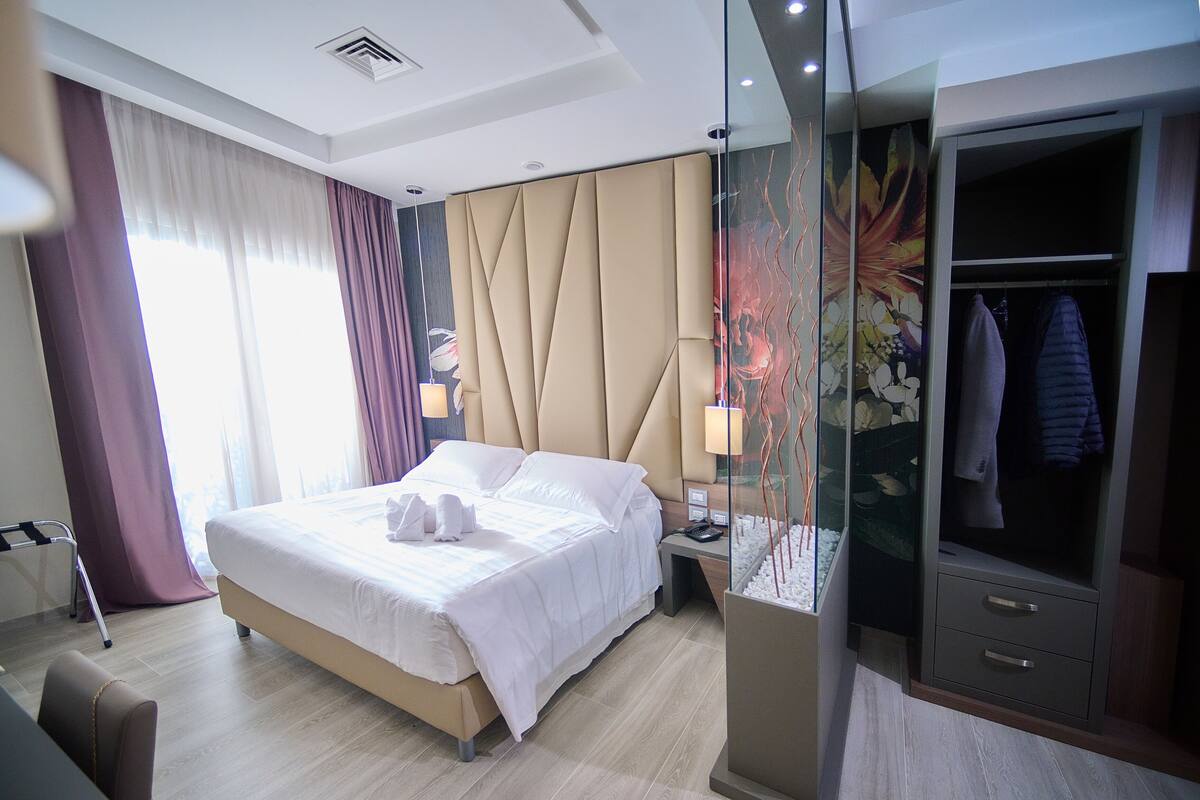 A modern hotel room with a large bed, a bedside table with a lamp, a wardrobe, and a glass partition with decorative elements.