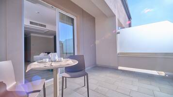 A balcony with a table and chairs, a sliding door leading to a bedroom, and a view of the sky.