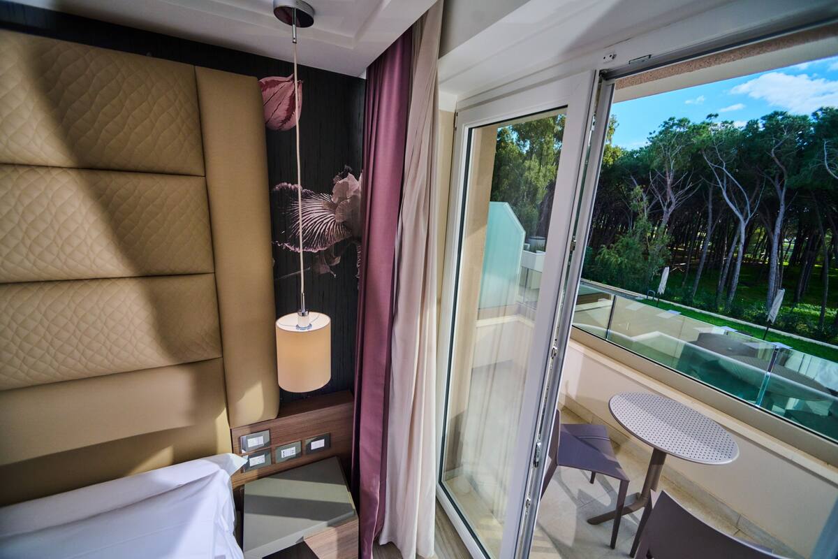 A hotel room with a large window, a balcony with a table and chairs, and a bed with a headboard.