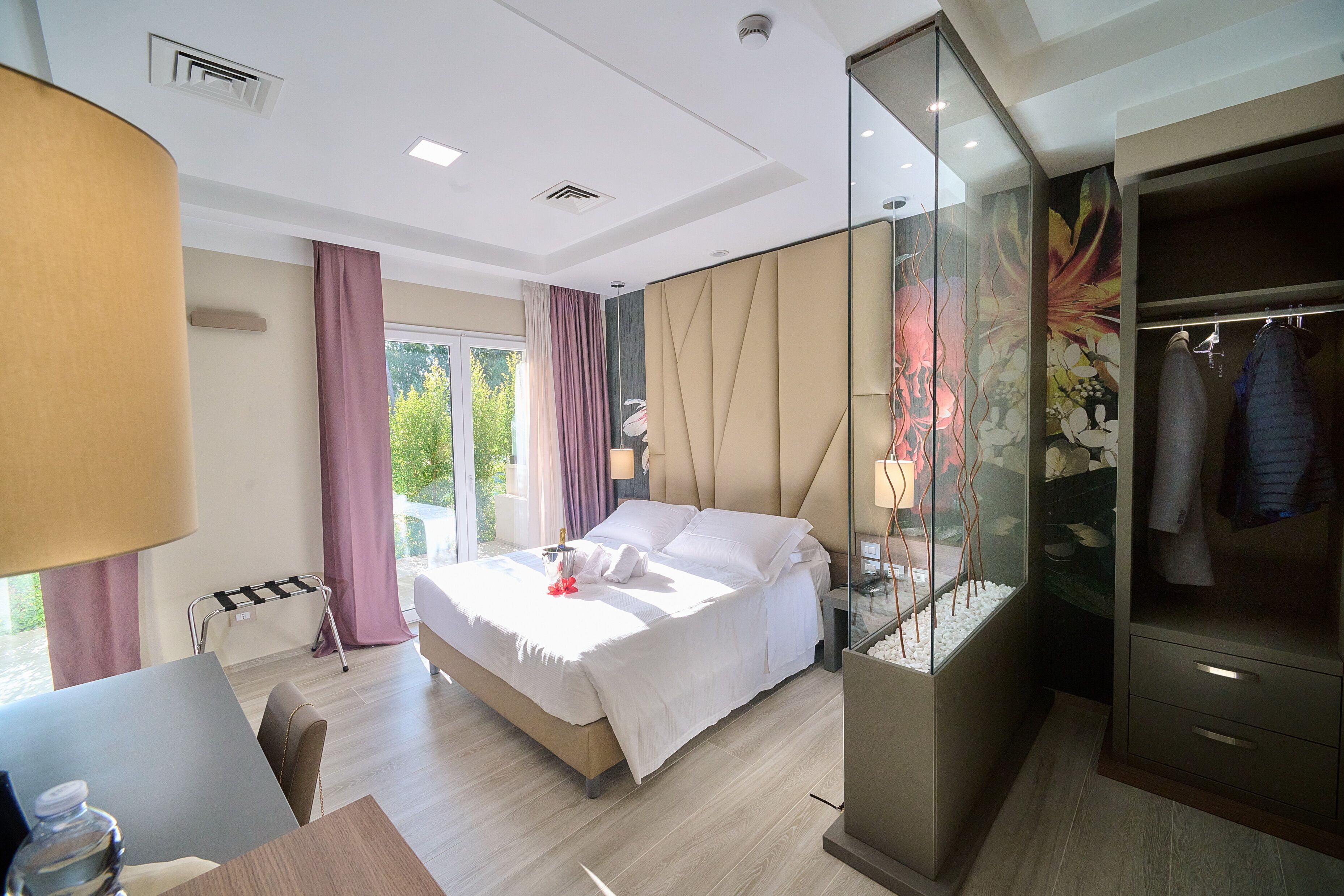 A modern hotel room with a large bed, a desk, a wardrobe, and a glass partition with a decorative fish tank.