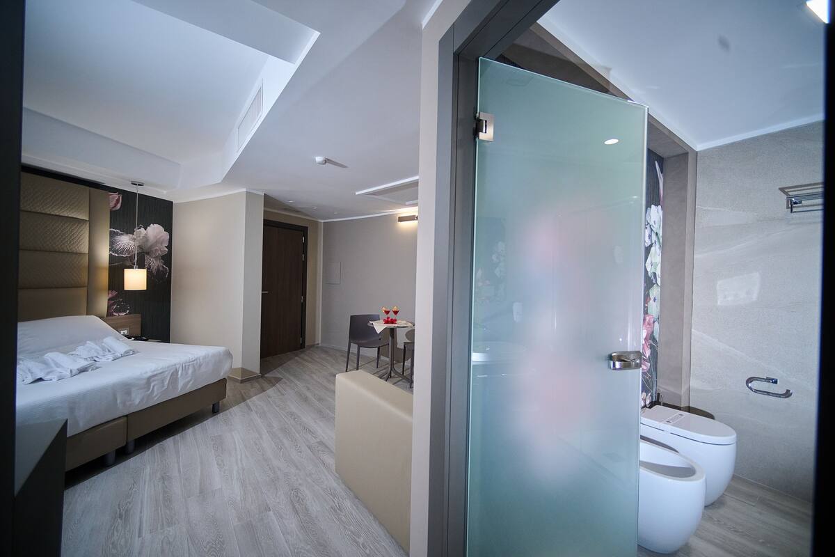 A modern hotel room with a bed, a small table with chairs, a glass-enclosed shower, and a toilet.