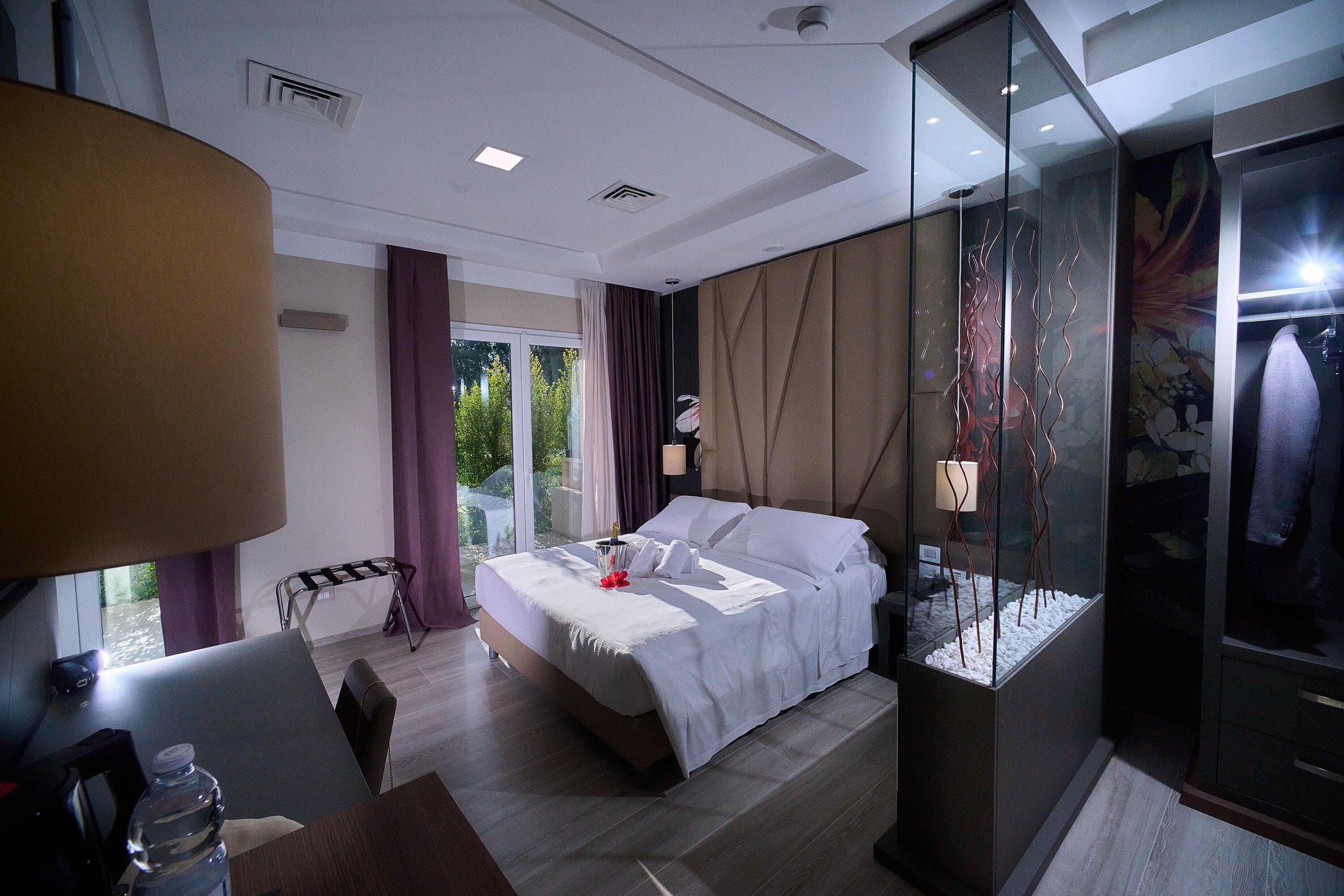 Een moderne hotelkamer met een groot bed, een bureau met stoel, een glazen scheiding en uitzicht op groen.