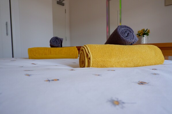 Una cama cuidadosamente tendida con toallas enrolladas en amarillo y azul oscuro, un florero con flores amarillas y una mesita de noche de madera.