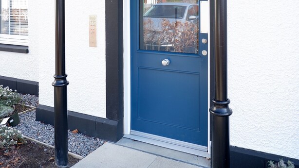 Una puerta azul con un pequeño ventanal, flanqueada por dos columnas negras, y una pared blanca con otro ventanal y una lámpara.