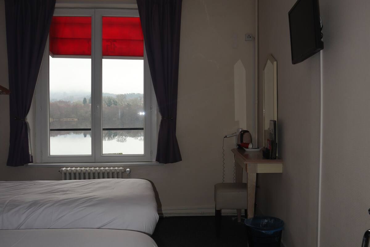 A bedroom with a bed, a window with curtains, a radiator, a desk with a mirror, a television, and a trash bin.