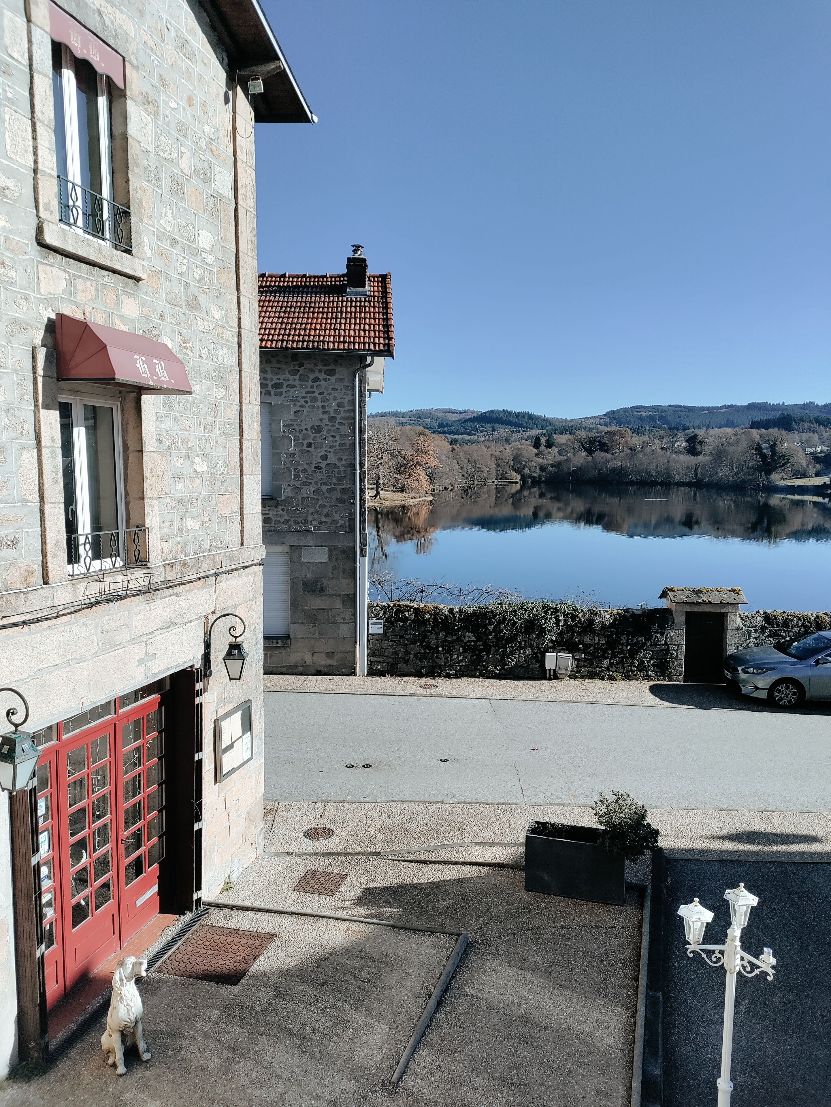 Un bâtiment en pierre avec une porte rouge, un petit chien devant, et une vue sur un lac et des collines en arrière-plan.