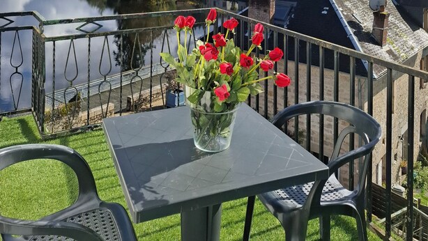 Un balcón con una mesa y sillas, un florero con flores rojas, y una vista a un lago y casas a lo lejos.