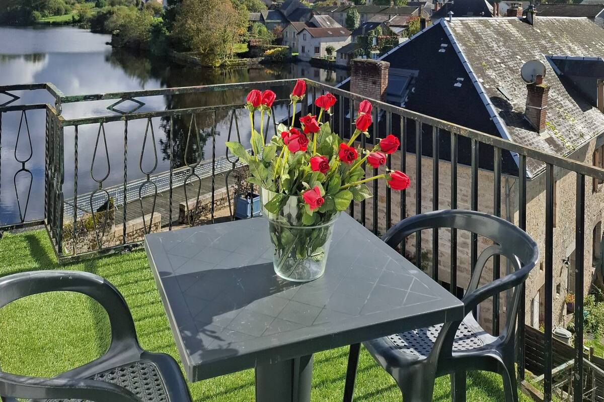 A balcony with a table and chairs, a vase of red flowers, and a view of a lake and houses in the distance.