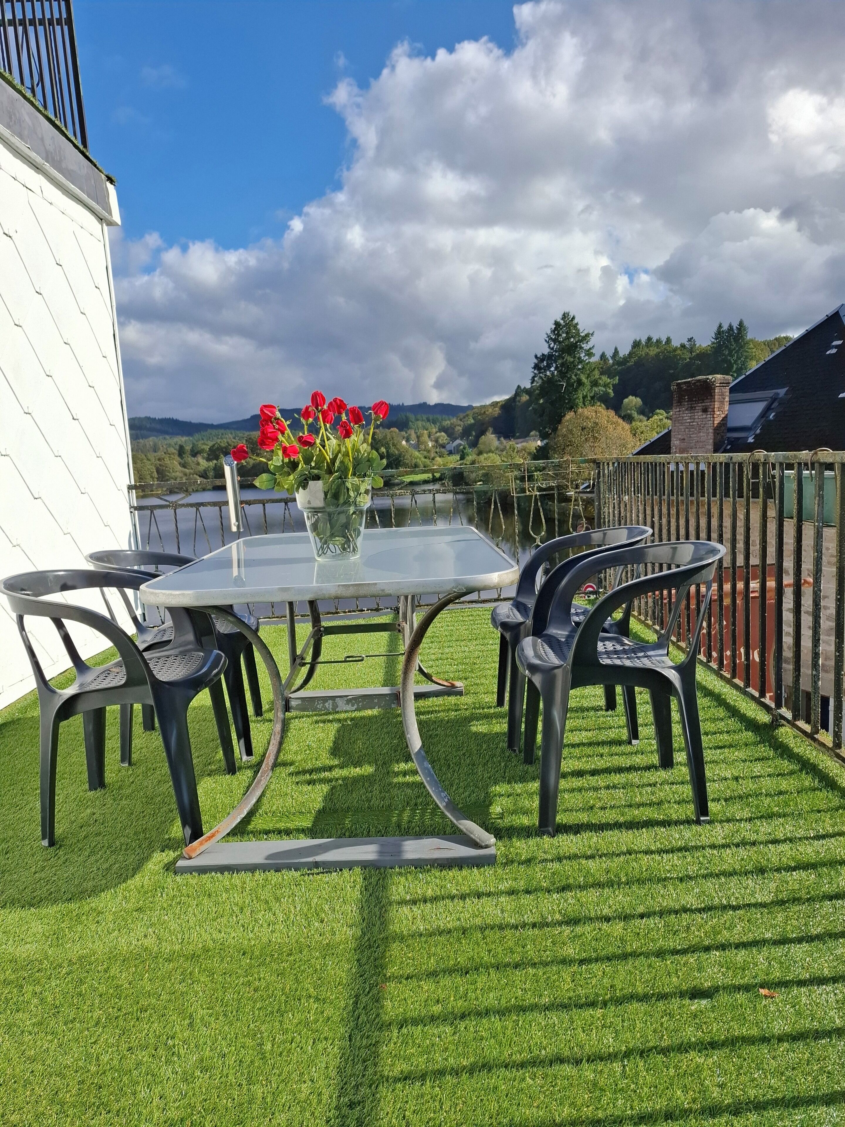 Mesa de vidrio con sillas y un jarrón de flores rojas sobre una terraza de césped con vista a un lago y colinas.