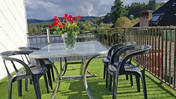 Mesa de vidrio con sillas y un jarrón de flores rojas sobre una terraza de césped con vista a un lago y colinas.