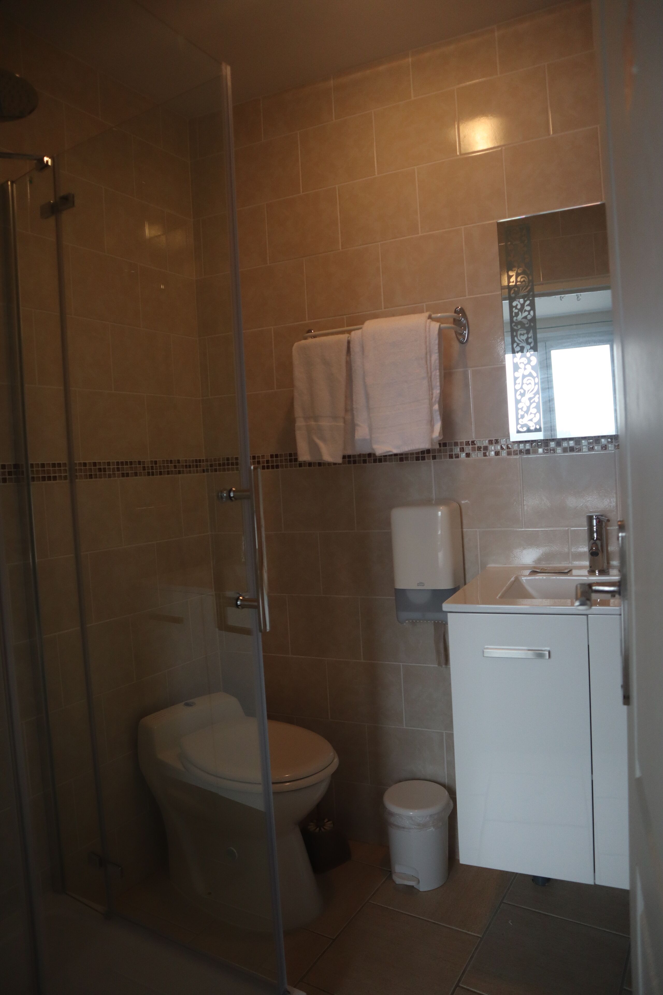 Un baño con ducha separada por mampara de vidrio, inodoro, lavamanos y un cubo de basura.
