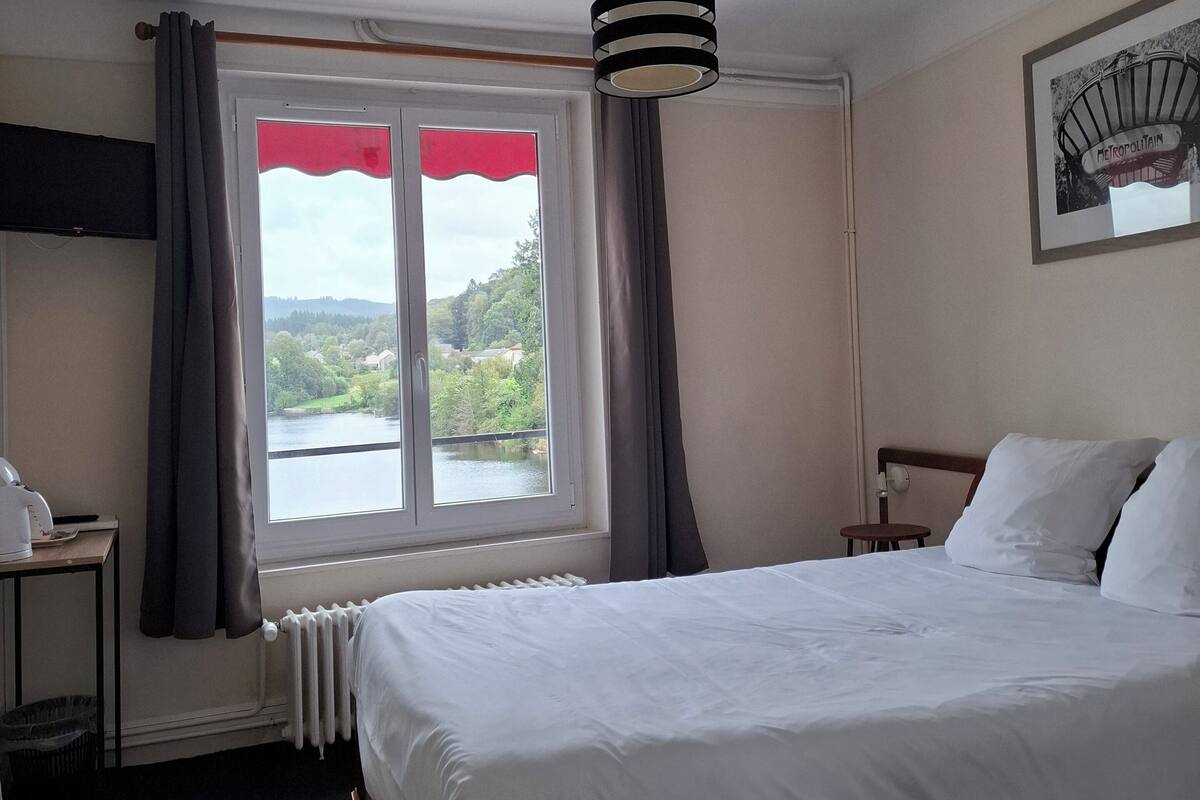A hotel room with a bed, a window with curtains, a radiator, a small table with a phone, and a picture on the wall.