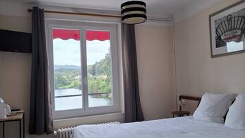 Een hotelkamer met een bed, een raam met gordijnen, een radiator, een klein tafeltje met een telefoon en een foto aan de muur.