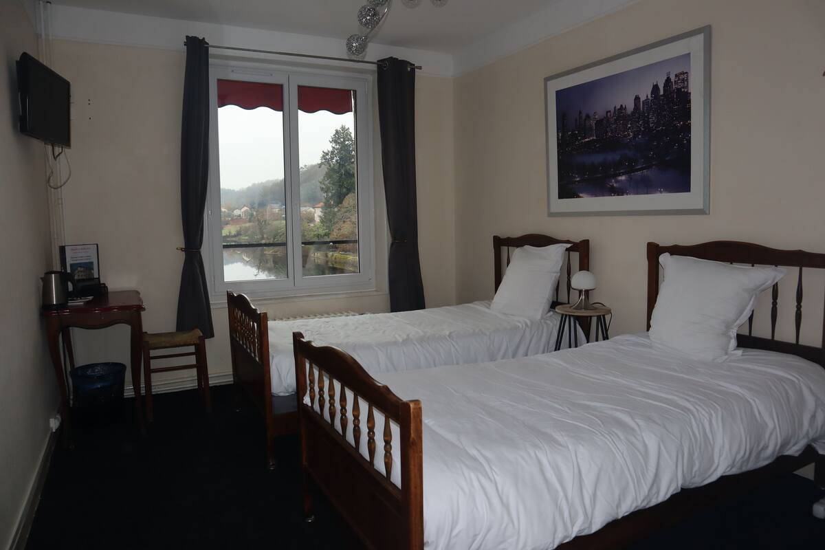 A hotel room with two single beds, a desk with a chair, a small table, a television, a window with curtains, and a cityscape painting on the wall.