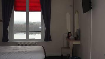 A bedroom with a large bed, a window with red curtains, a radiator, a desk with a mirror, a television, and a trash bin.