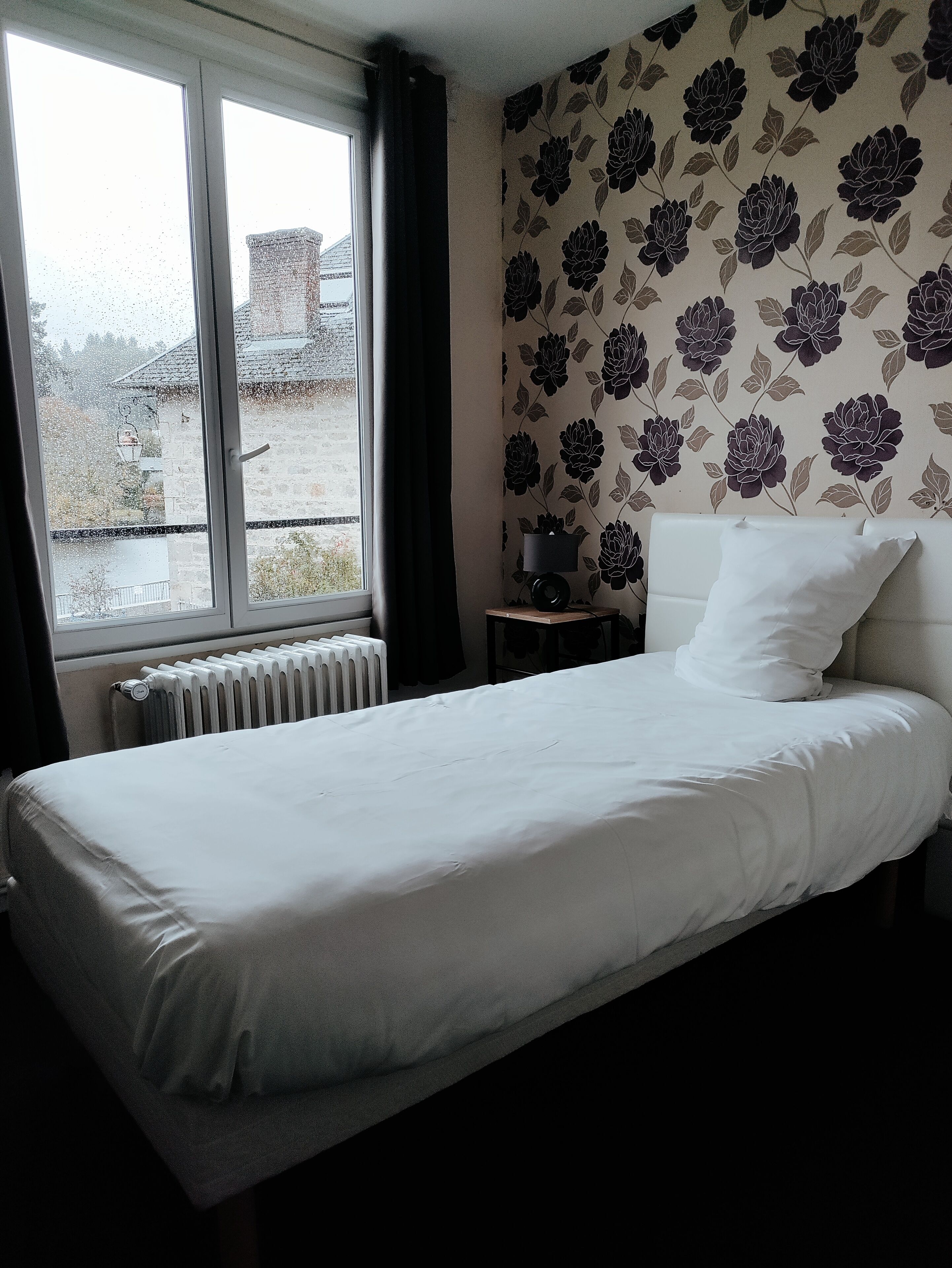 A bedroom with a bed, a radiator, a window with curtains, and a wall with a floral pattern.