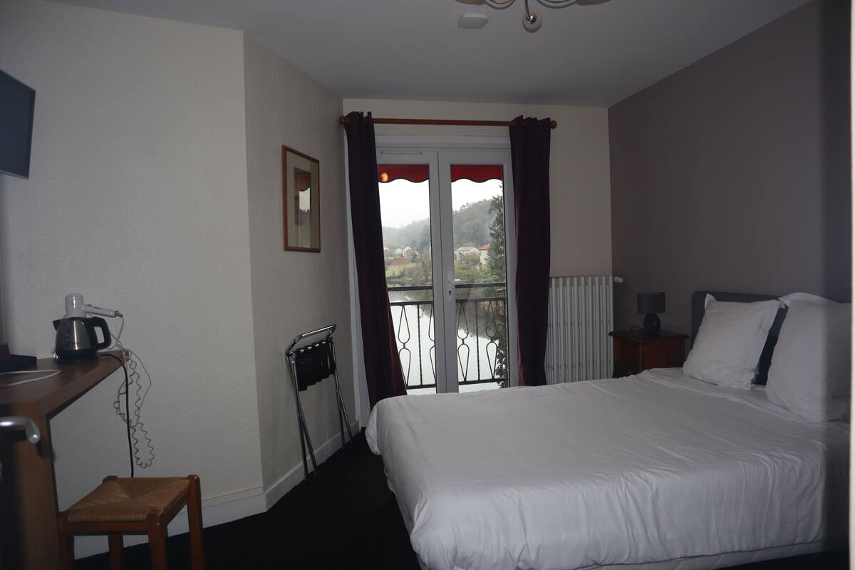 A hotel room with a bed, a desk with a kettle, a chair, a radiator, and a window with curtains.