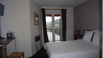 A hotel room with a bed, a desk with a kettle, a chair, a radiator, and a window with curtains.