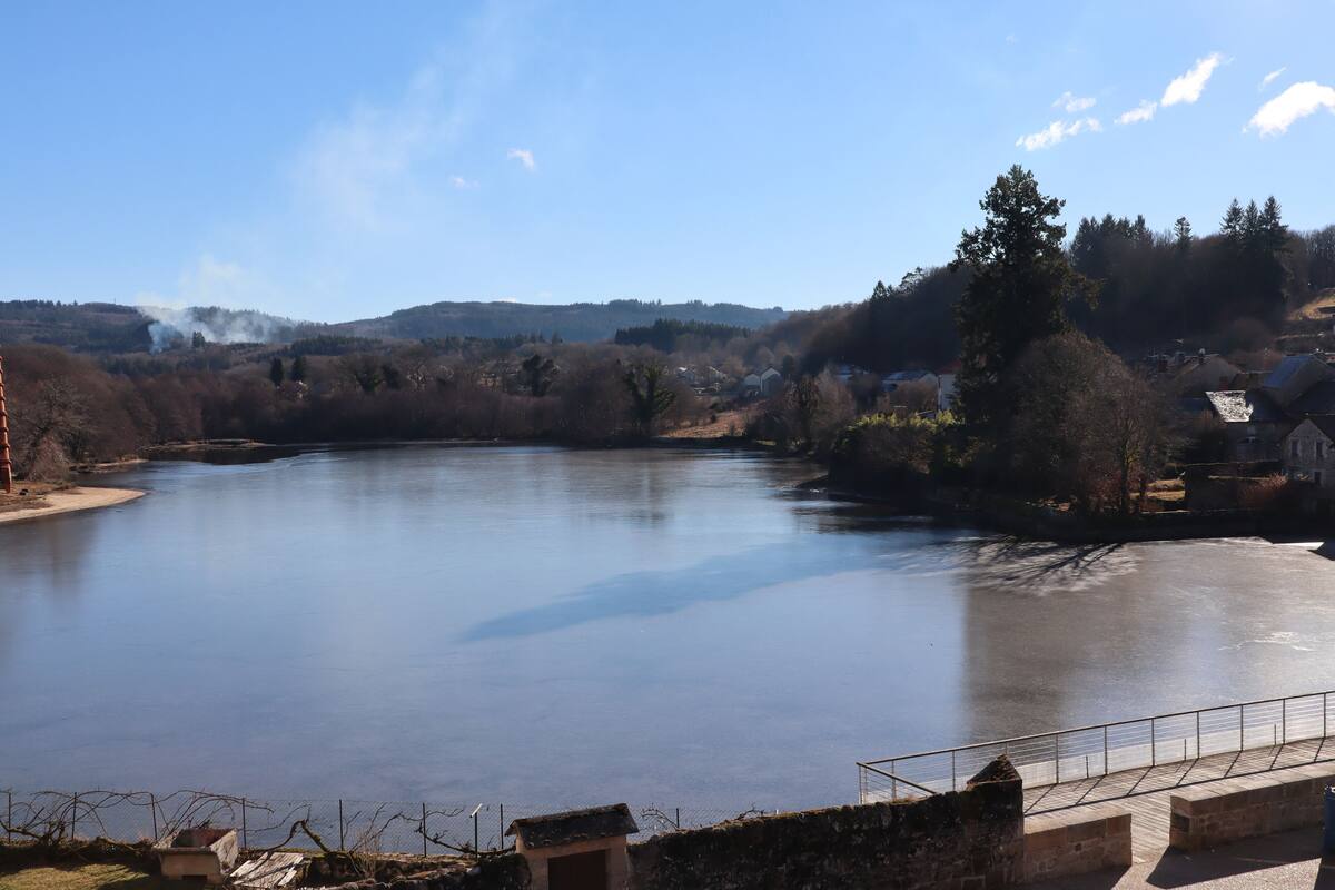 A calm river with a stone bridge in the foreground and a hilly landscape in the background.