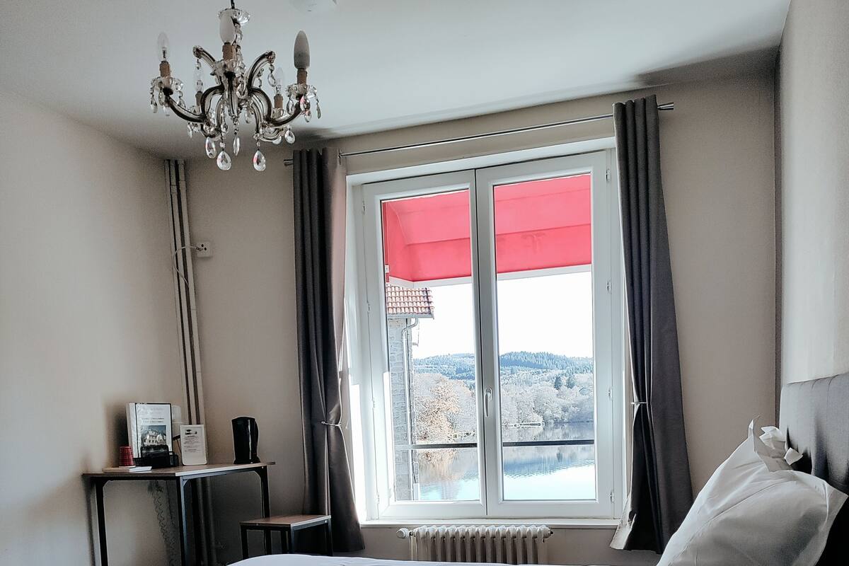 A hotel room with a bed, a desk, a radiator, a window with curtains, and a chandelier.