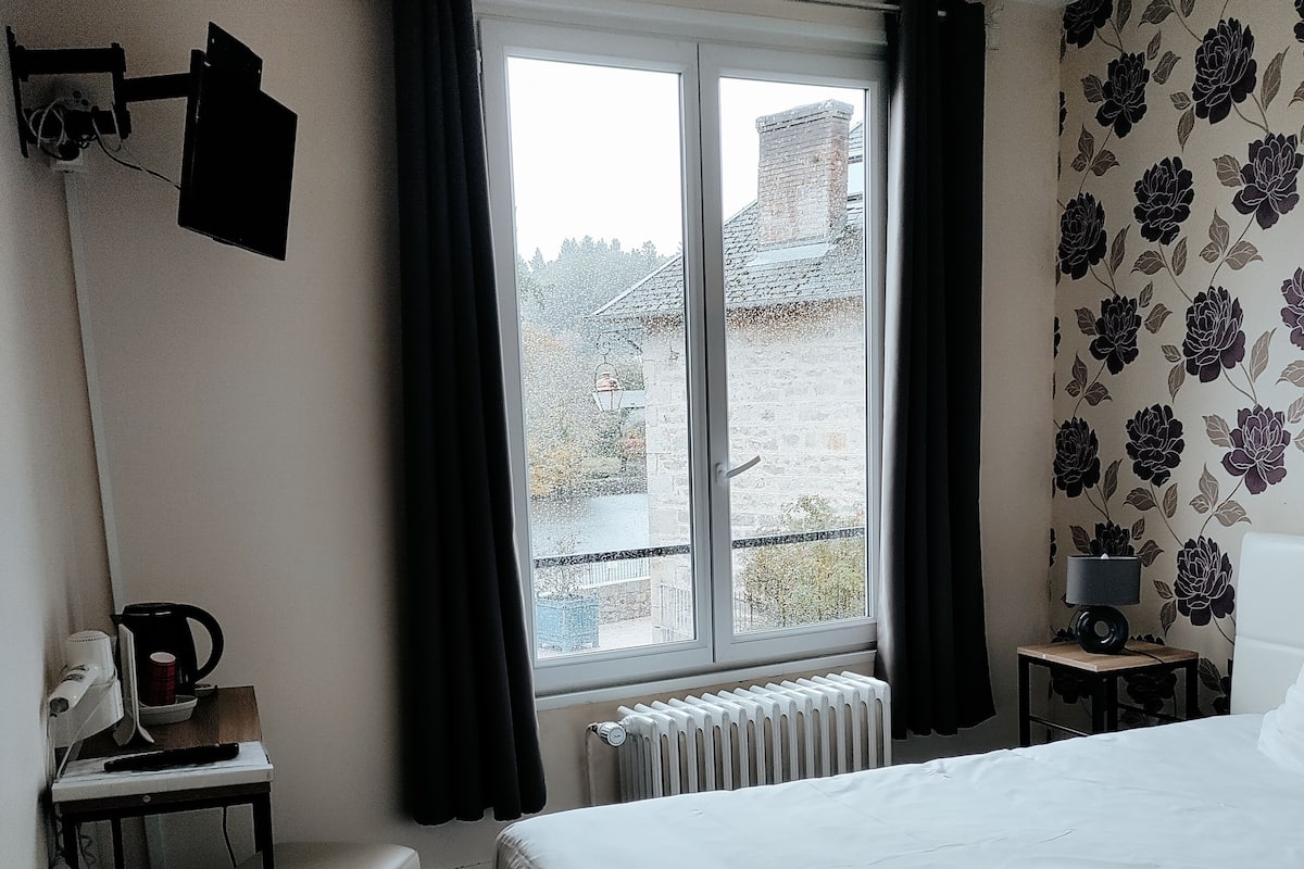 A hotel room with a bed, a desk with a lamp, a radiator, and a window with curtains.