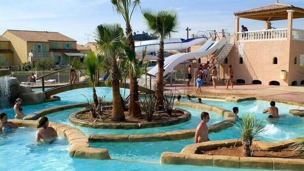 Un espace piscine doté de plusieurs éléments aquatiques, notamment un toboggan et une aire de jeux aquatique circulaire entourée de palmiers.