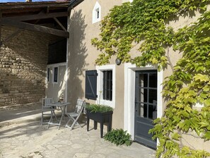 A stone building with a white door and windows, a small table with chairs, and potted plants.