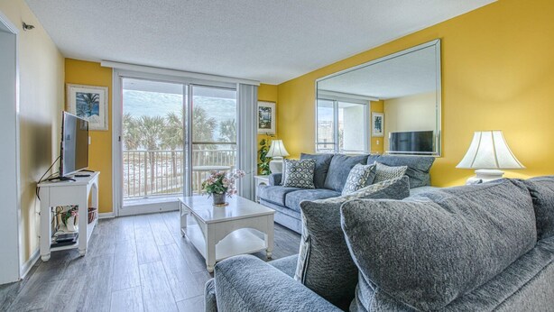 A living room with a grey sofa, a coffee table, and a TV on a stand. There is a balcony with a view of palm trees. - Updated Beach Front Property!!! (Destin)