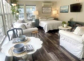 A modern hotel room with a large bed, two white sofas, a dining table with bowls, a small round table, and a TV on the wall.