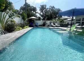 A rectangular swimming pool with clear blue water, surrounded by a white fence and lush greenery. There are lounge chairs and umbrellas providing shade.